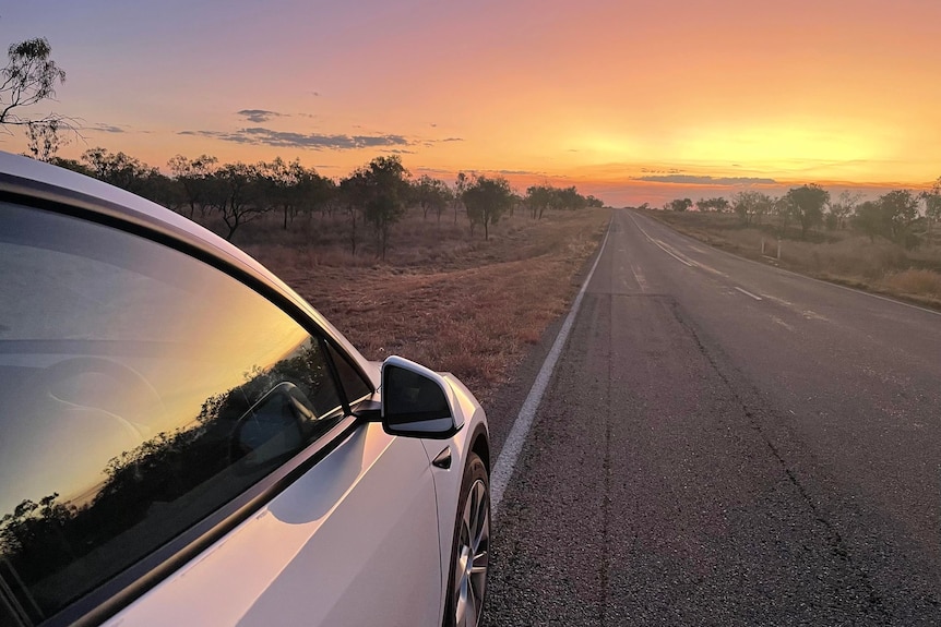An electric vehicle parked on the highway with a peach sunset in the horizon