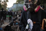 A student wears a gas mask and holds a molotov cocktail as groups of students gather in the background.