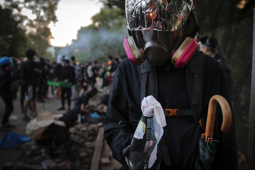 A student wears a gas mask and holds a molotov cocktail as groups of students gather in the background.