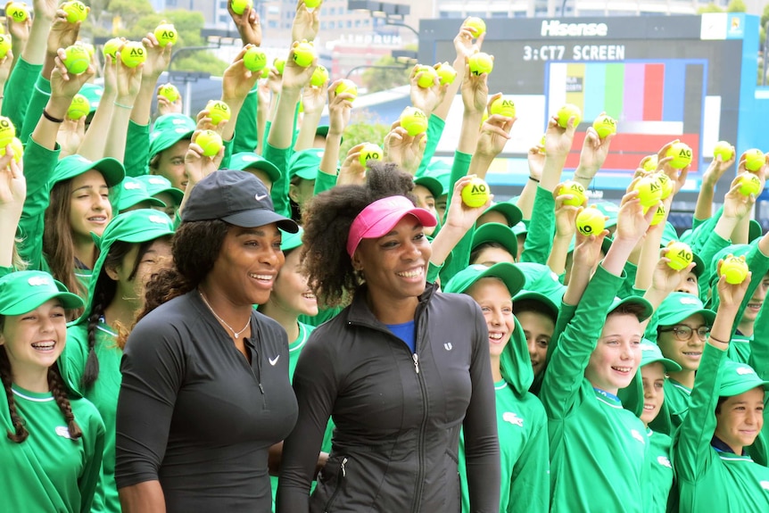 Serena and Venus posing in front of a bunch of kids wearing green and holding up tennis balls