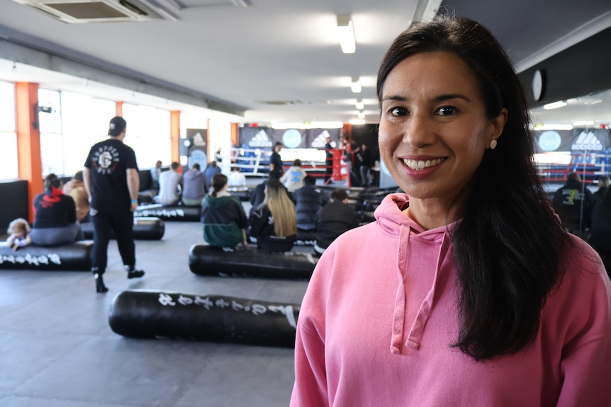 A woman in a pink hoodie smiles, standing in a boxing gym.