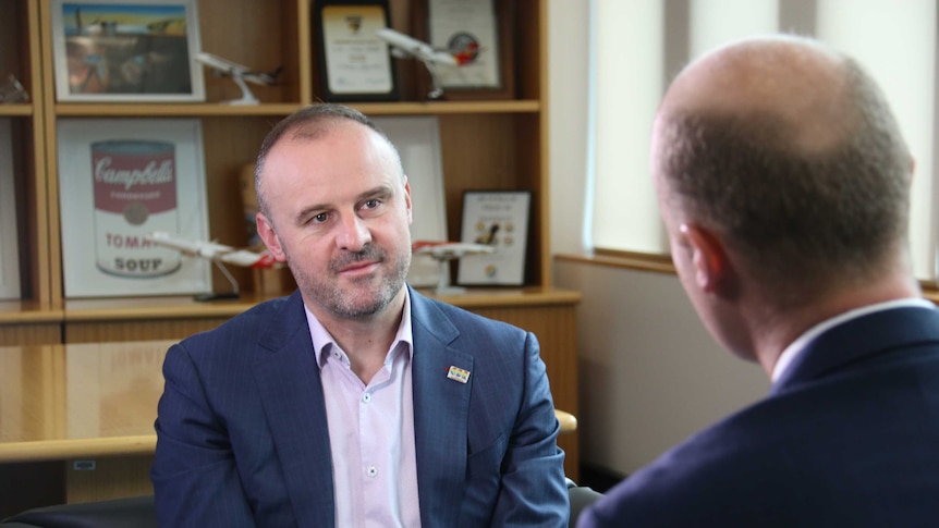 A man in  a suit sits in a chair in his office, speaking to a journalist whose back is to the camera.