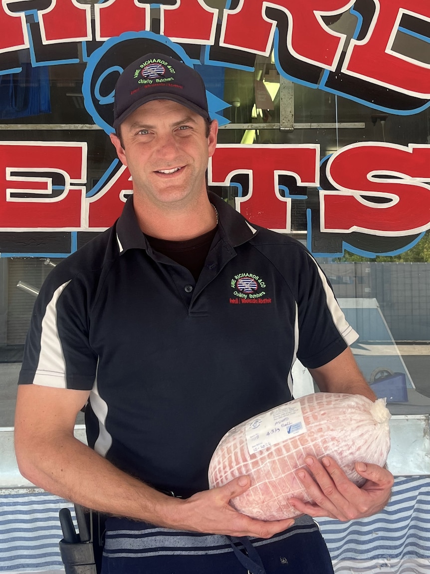 A man smiles at the camera while holding turkey meat in front of a butcher store. 