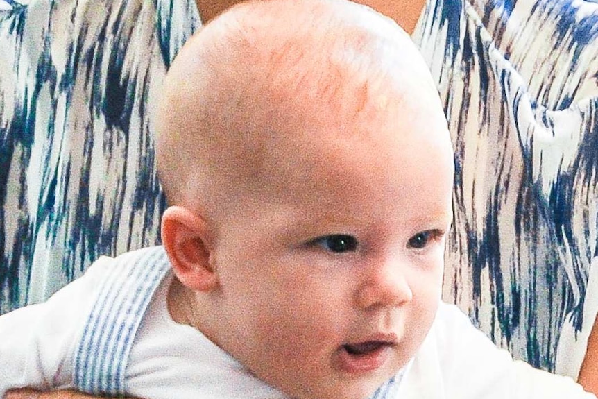 Blonde-haired baby boy wearing white, long-sleeved top and blue and white striped overalls, held by his mother