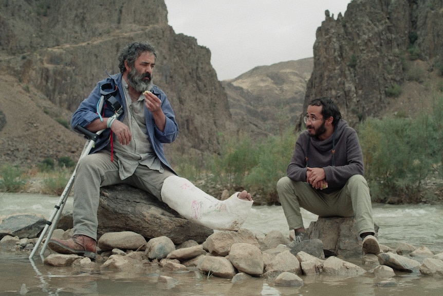 A father and adult son are sitting chatting on some rocks by a river with dry rocky mountains in the background. 