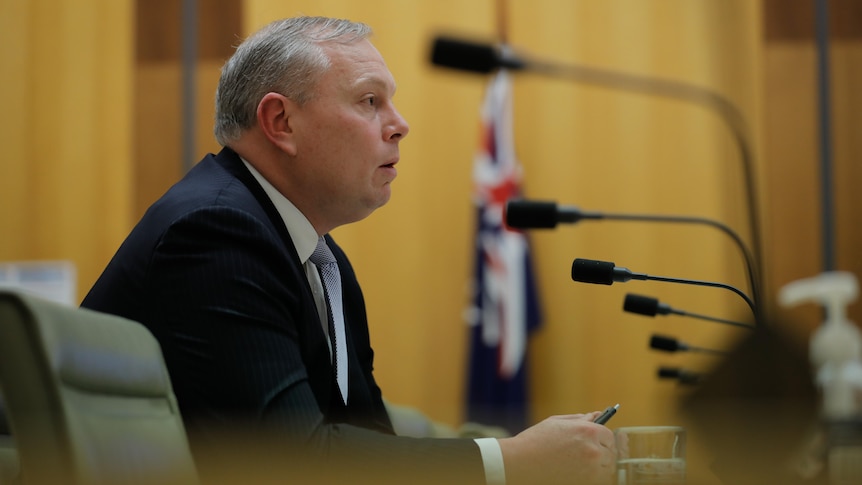 A man sitting at a table facing several microphones with an Australian flag in the background.
