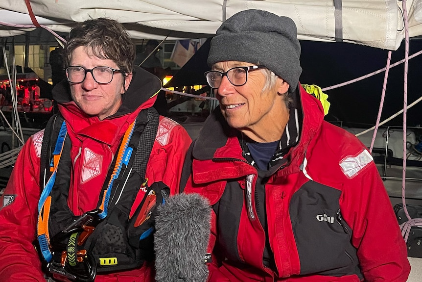 Two women being interviewed. They are wearing glasses and red, weather-proof jackets. Kathy has a grey beanie on her head.