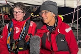 Two women being interviewed. They are wearing glasses and red, weather-proof jackets. Kathy has a grey beanie on her head.