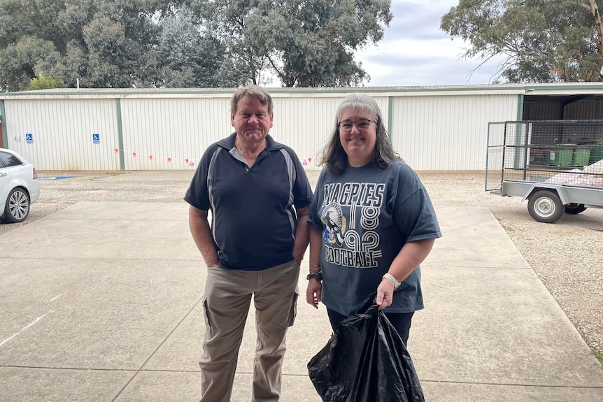 A man and woman stand smiling outside, holding a garbage bag