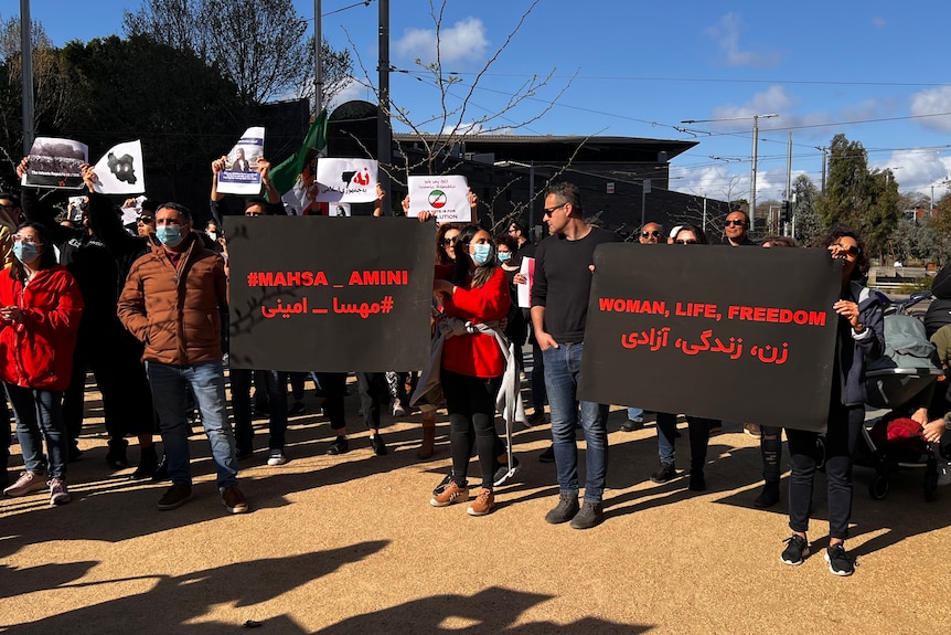 A group of people stand around with signs reading "Woman, Life, Freedom".