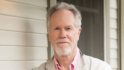 Loudon Wainwright III wearing a pink shirt and cream jacket leaning on a weatherboard wall