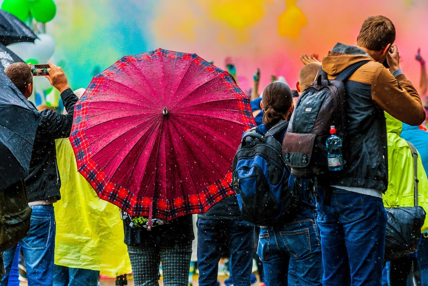 Young people stand in the rain at a festival, with one person holding a large pink umbrella. In the background.