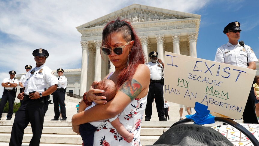 A woman holds her baby daughter on the Supreme Court steps next to a sign that says "I exist because my Mom had an abortion"