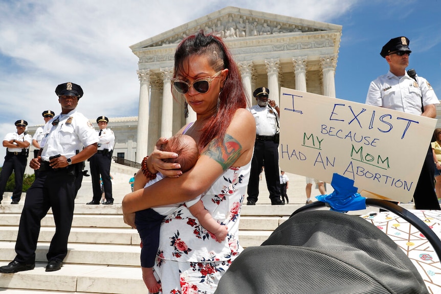 A woman holds her baby daughter on the Supreme Court steps next to a sign that says "I exist because my Mom had an abortion"