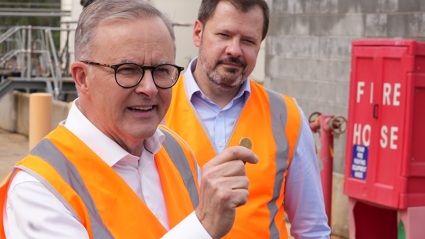 Two men in orange hi vis vest, with one holding up a dollar coin