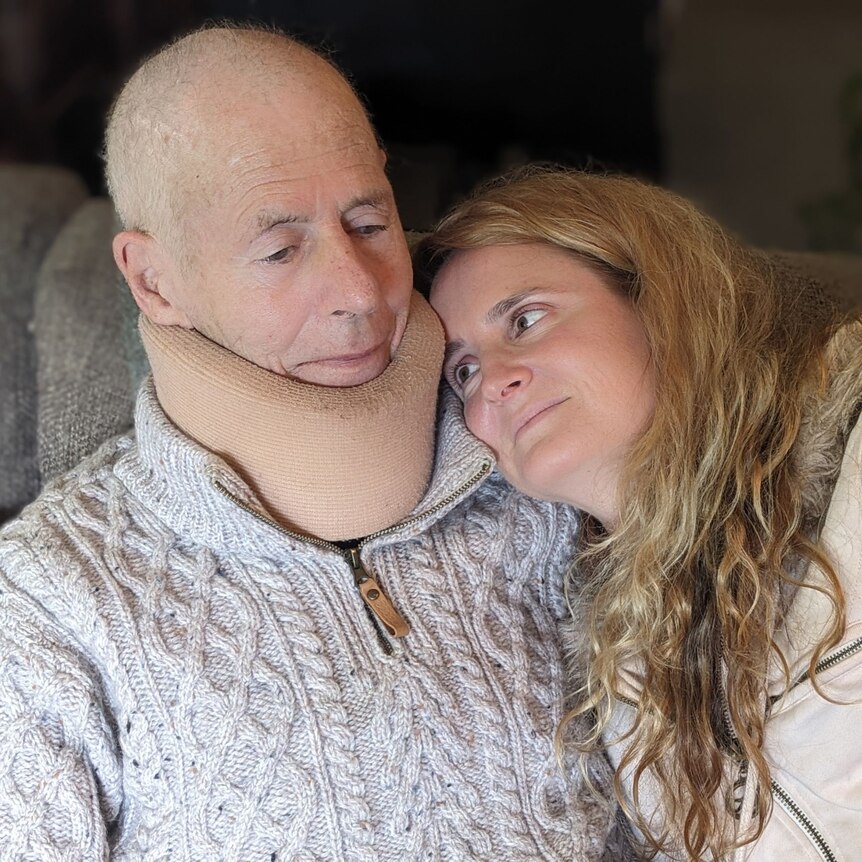 A man in a neck brace lying against cushions, a women rests her head on his shoulder, looking up at him