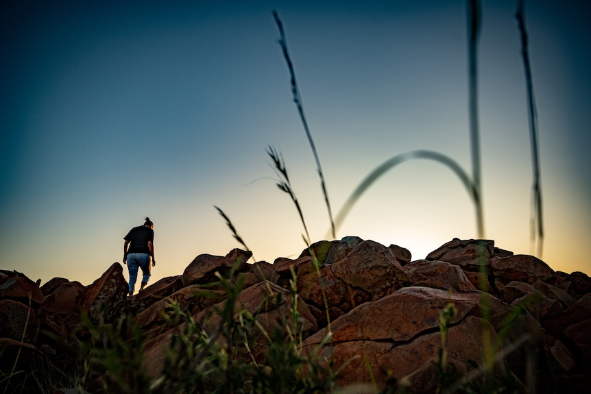 The sun is rising over rocks in Murujuga, back to camera, Raelene Cooper makes her way to the top.