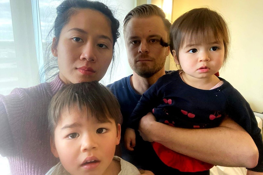 A family portrait taken in a hotel room of a woman wearing a purple jumper, a man wearing a navy t-shirt, and two children.