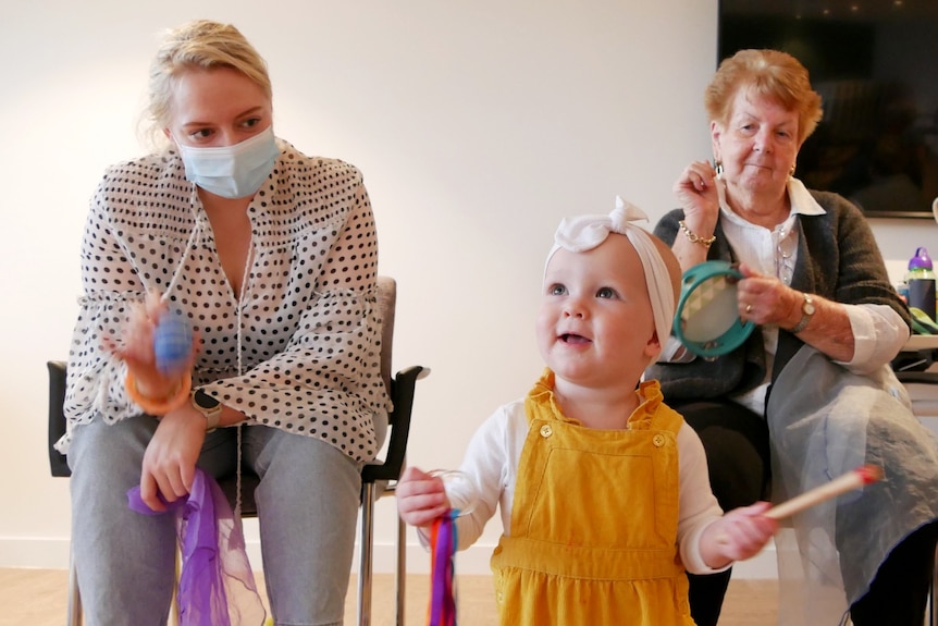 A small baby stands in front of two women sitting in chairs. 