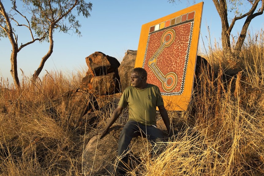 Man sits on grassy hill with Indigenous painting behind him