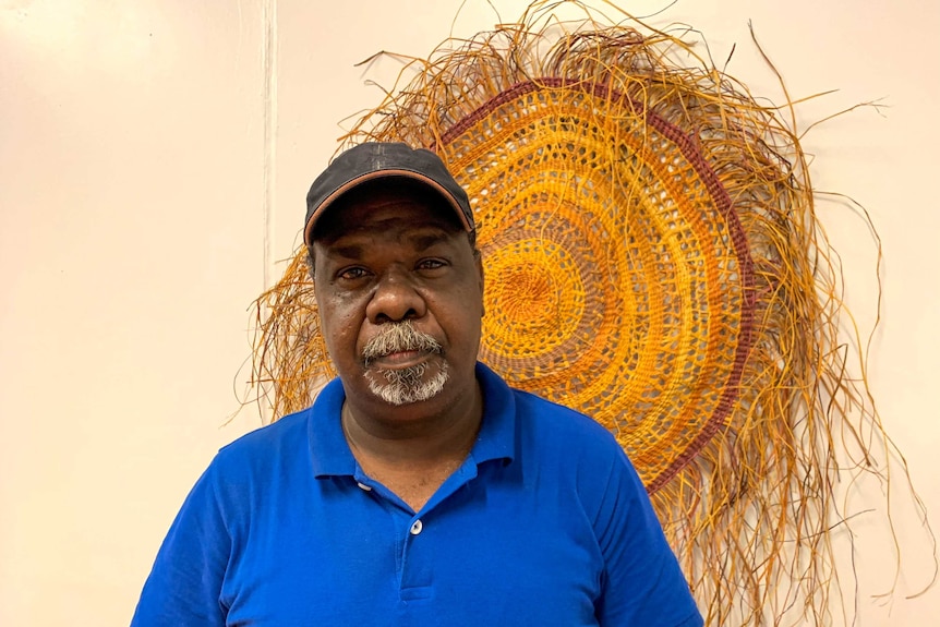 An Indigenous man stands next to a piece of artwork, he is wearing a cap and is looking directly into the camera.