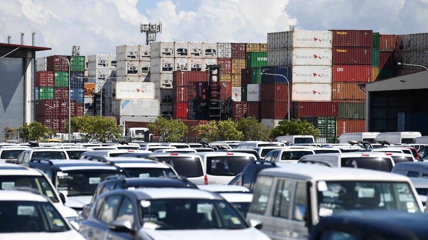Rows of cars in a car park, with stacked shipping containers behind them.