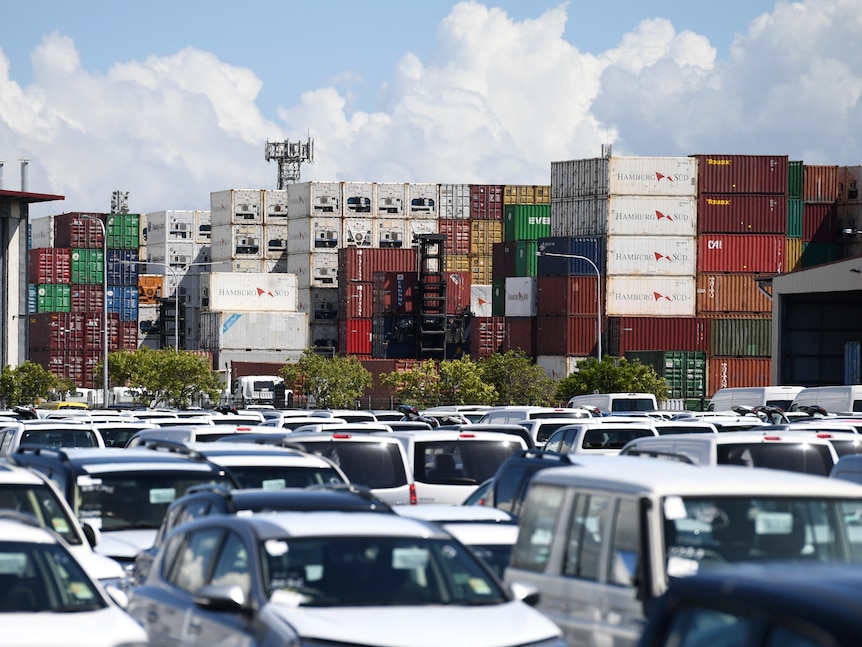 Rows of cars in a car park, with stacked shipping containers behind them.