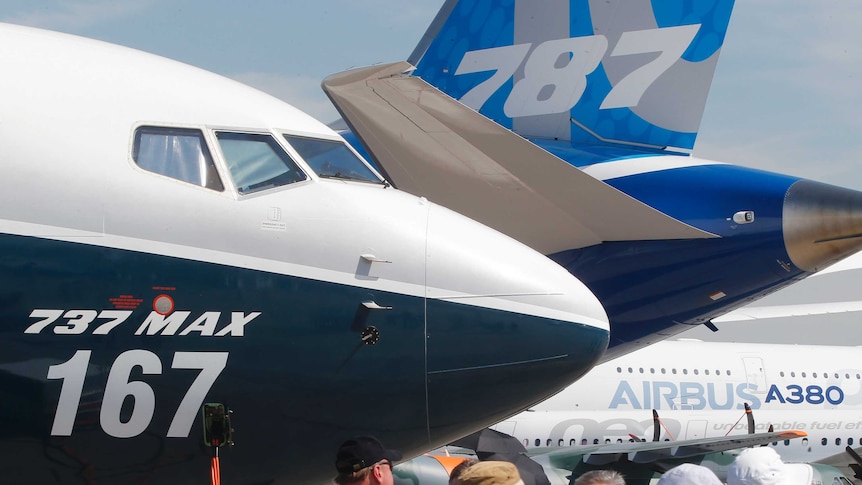 A Boeing 737 MAX nose is shown close-up, with the tail of a Boeing 787 and the fuselage of an Airbus A380 behind it.