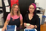 Two women sitting at a desk. One of them has bright pink heair and is holding a cup of tea. They have copies of the QLRC report.