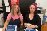 Two women sitting at a desk. One of them has bright pink heair and is holding a cup of tea. They have copies of the QLRC report.