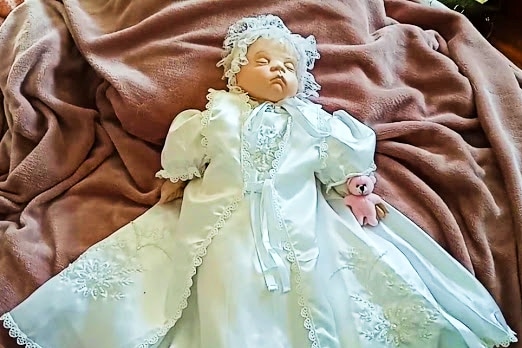 A porcelain doll in a white christening dress and bonnet lies on a bed. 