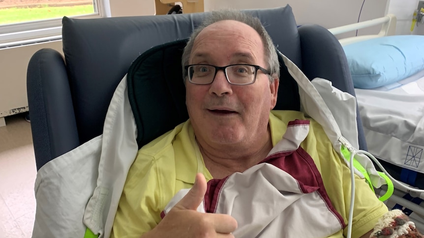 A man sitting in a chair in a hospital room giving a thumbs up and smiling.