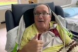 A man sitting in a chair in a hospital room giving a thumbs up and smiling.