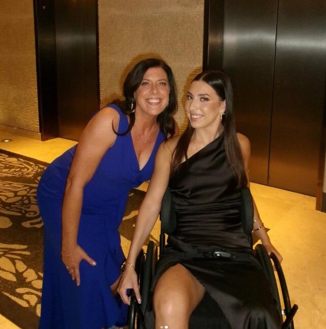 Uma mulher sorri para a câmera ao lado de outra mulher em uma cadeira de rodas.