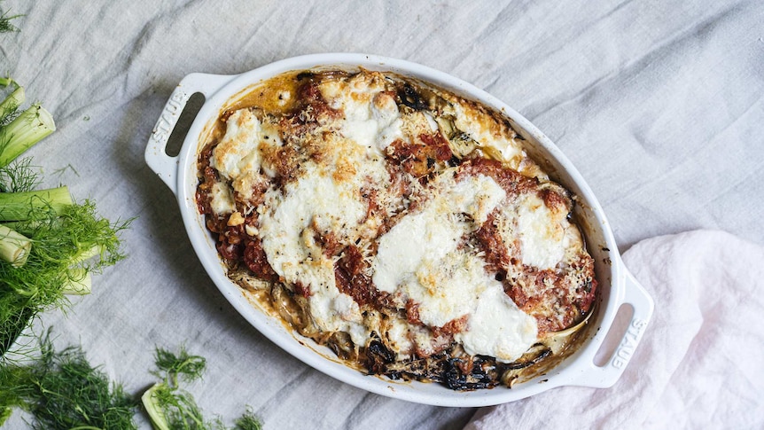 A baking dish of fennel parmigiana sits on a linen tablecloth with fennel fronds in the corner, a vegetarian family dinner.