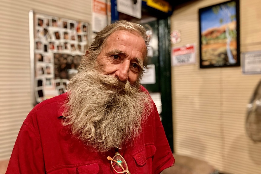 A man with a big grey beard and red shirt looks at the camera