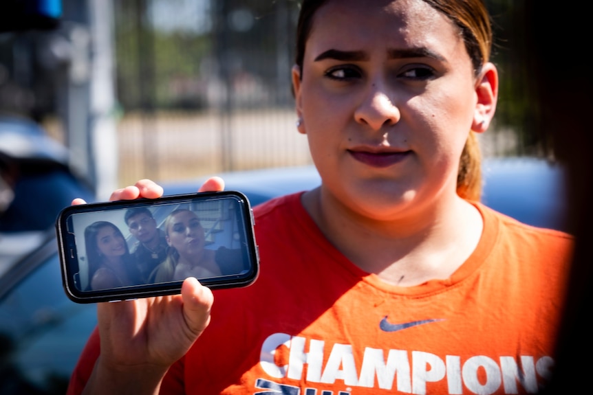 A woman in an orange shirt holds up a photo on her phone of herself with two friends, one being the victim.
