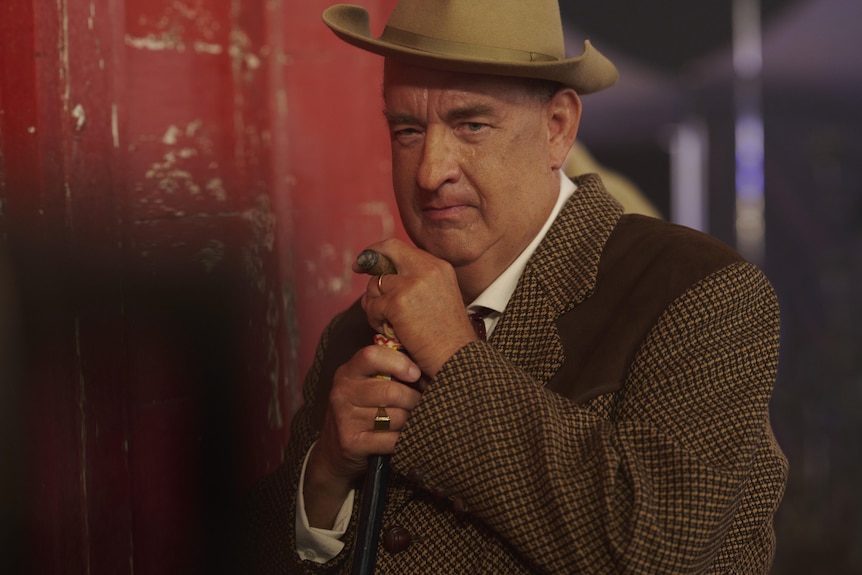 Tom Hanks in hat and clutches a walking stick while smoking a cigar and looking intently at something offscreen.