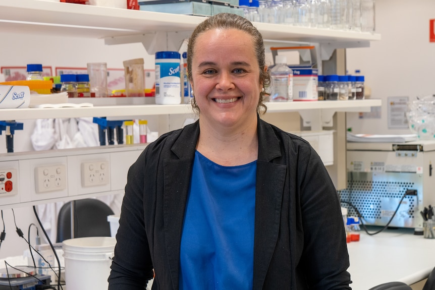 Katia Nones in blue top, black jacket, curly brown hair pulled back in a lab in front of shelves of medical tools, supplies