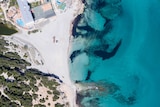 You view an aerial photo of a wide, sandy beach next to azure blue waters.