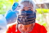 An older woman in a face mask and red t-shirts gets her temperature scanned