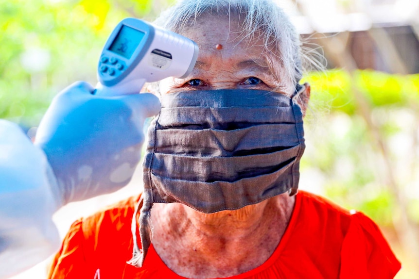 An older woman in a face mask and red t-shirts gets her temperature scanned