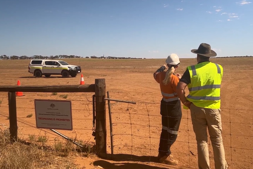 A man and woman in high visibility clothes and hardhats point across a barren, dry paddock