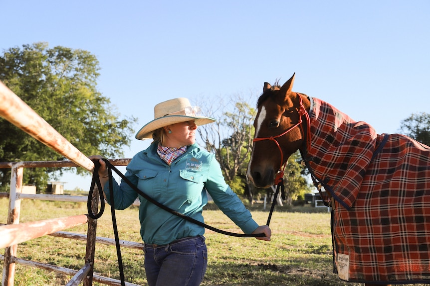 Woman wears brow-brimmed hat, jeans, long-sleeve shirt and neckerchief as she ties a horse wearing a blanket up to a fence