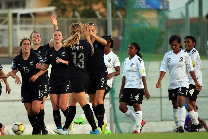 New Zealand female players in black shirts high five as scored a goal and Fijian players in white shirts look on.
