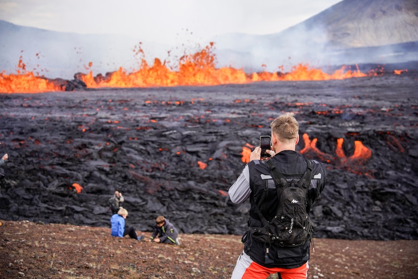 A man takes a photo on his phone as two other people sit in front of a mass of black and red lava. Lava explodes on horizon