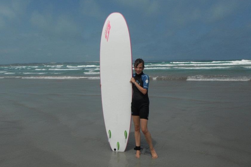 Young Chloé Hayden stands next to a long surfboard on a beach. 