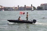 Two shirtless men aboard a motorboat in Venice with a flag that reads "no big ships" in Italian.