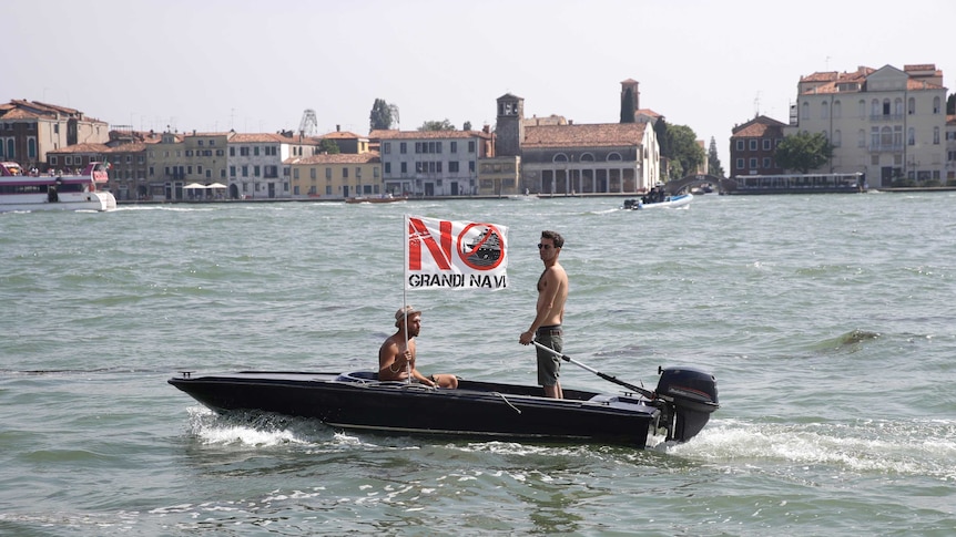 Two shirtless men aboard a motorboat in Venice with a flag that reads "no big ships" in Italian.