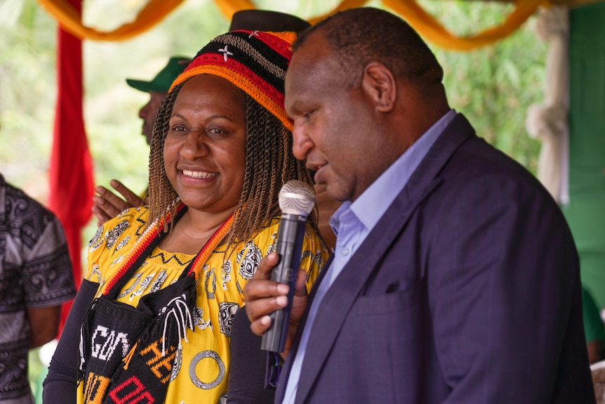 A woman wearing a beanie and a yellow top on stage stands next to a man speaking into a microphone.